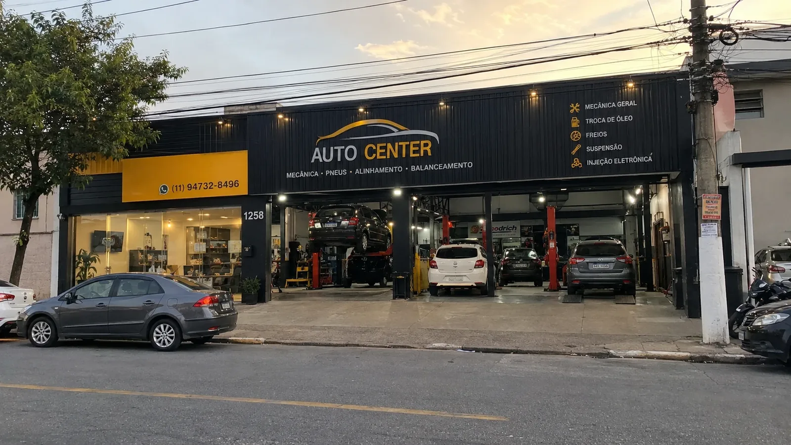 Fachada de um auto center no fim da tarde, com carros nos elevadores e a lista de serviços na placa